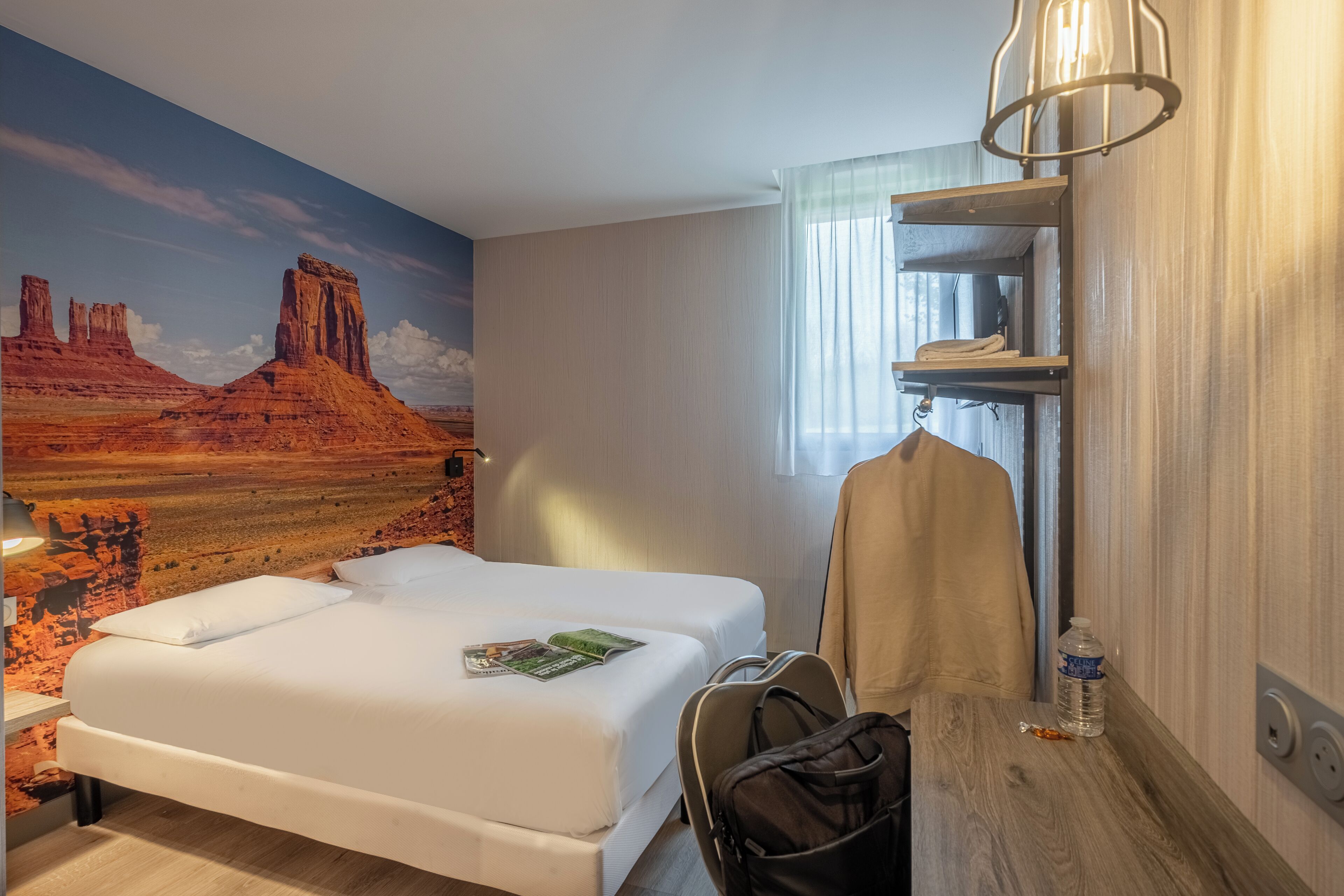 A hotel room with a large desert mural, a bed with white linens, a wooden nightstand, a hanging coat, and a water bottle.