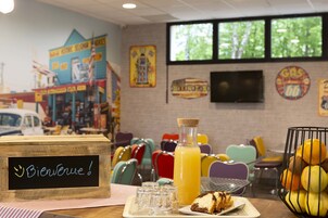 Un interior de restaurante con sillas coloridas, un letrero de madera con 'Bienvenue!', un vaso de jugo de naranja y un plato con una porción de pastel.