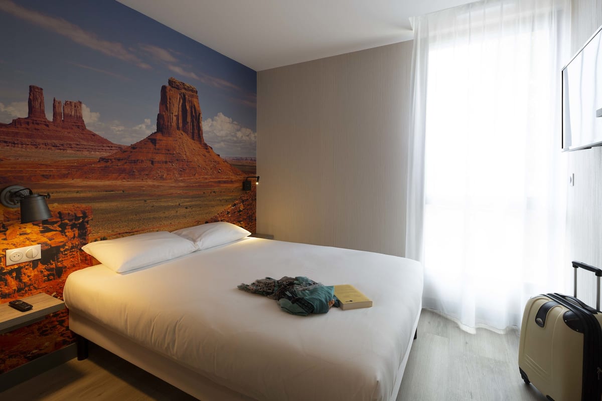 Hotelkamer met een bed, nachtkastje, lamp, koffer en een woestijnmotief op de muur.