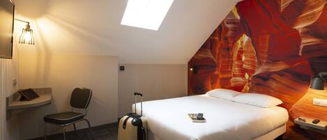 A hotel room with a bed, a desk, a chair, a suitcase, a skylight, and a large abstract mural on the wall.