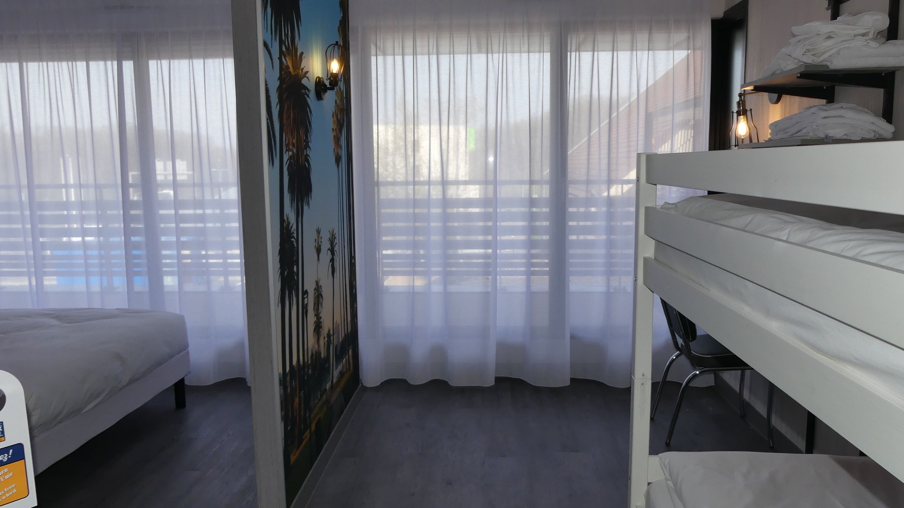 A hotel room with a bunk bed, a bed, a wall-mounted shelf with towels, and a window with sheer curtains.