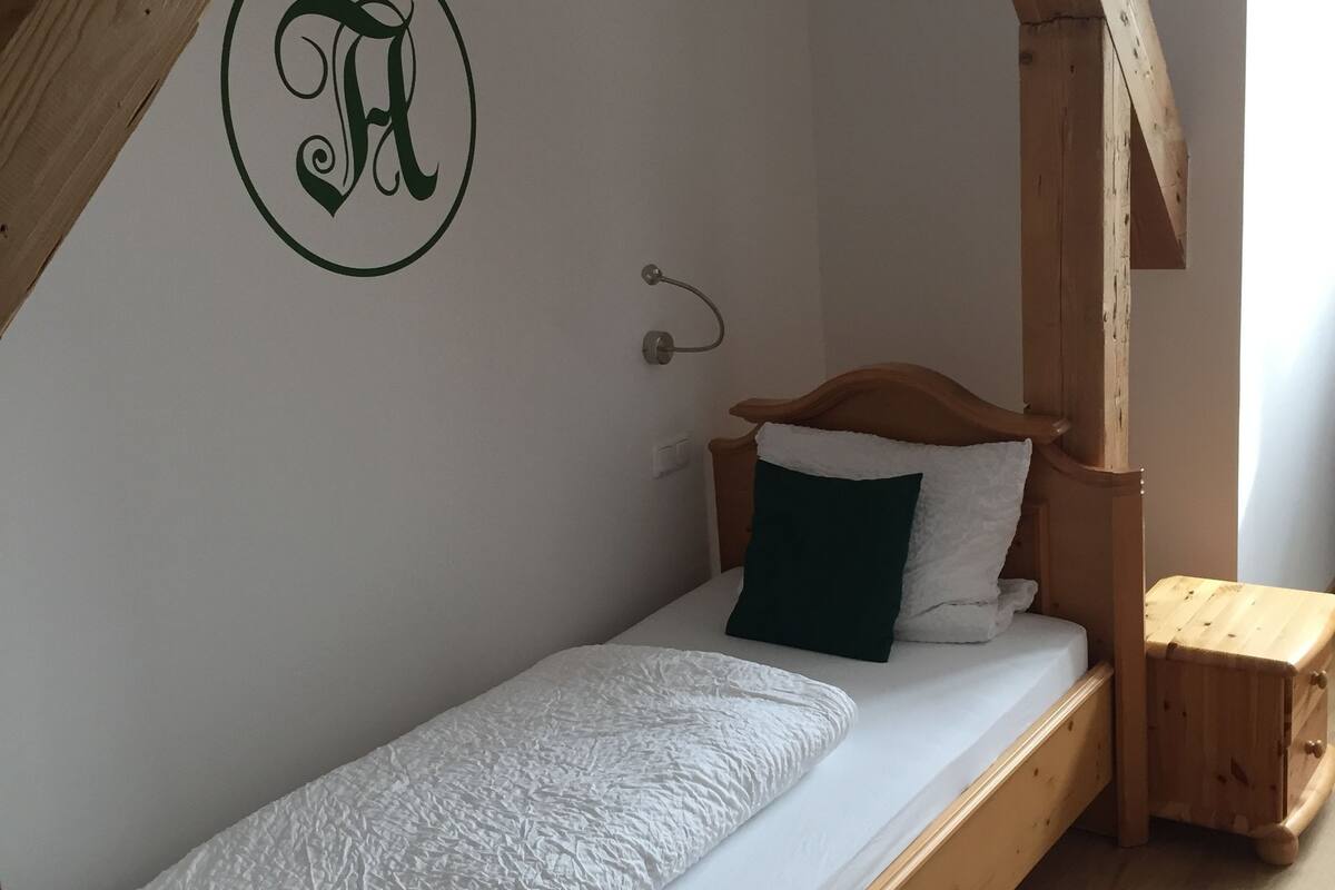 A single wooden bed with a white mattress and a green pillow in a room with exposed wooden beams and a white wall.