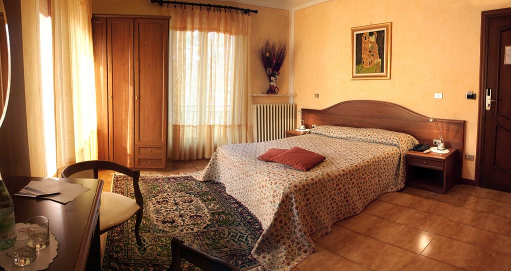 Un dormitorio con cama, escritorio, silla, radiador, ventana con cortinas y un cuadro en la pared.