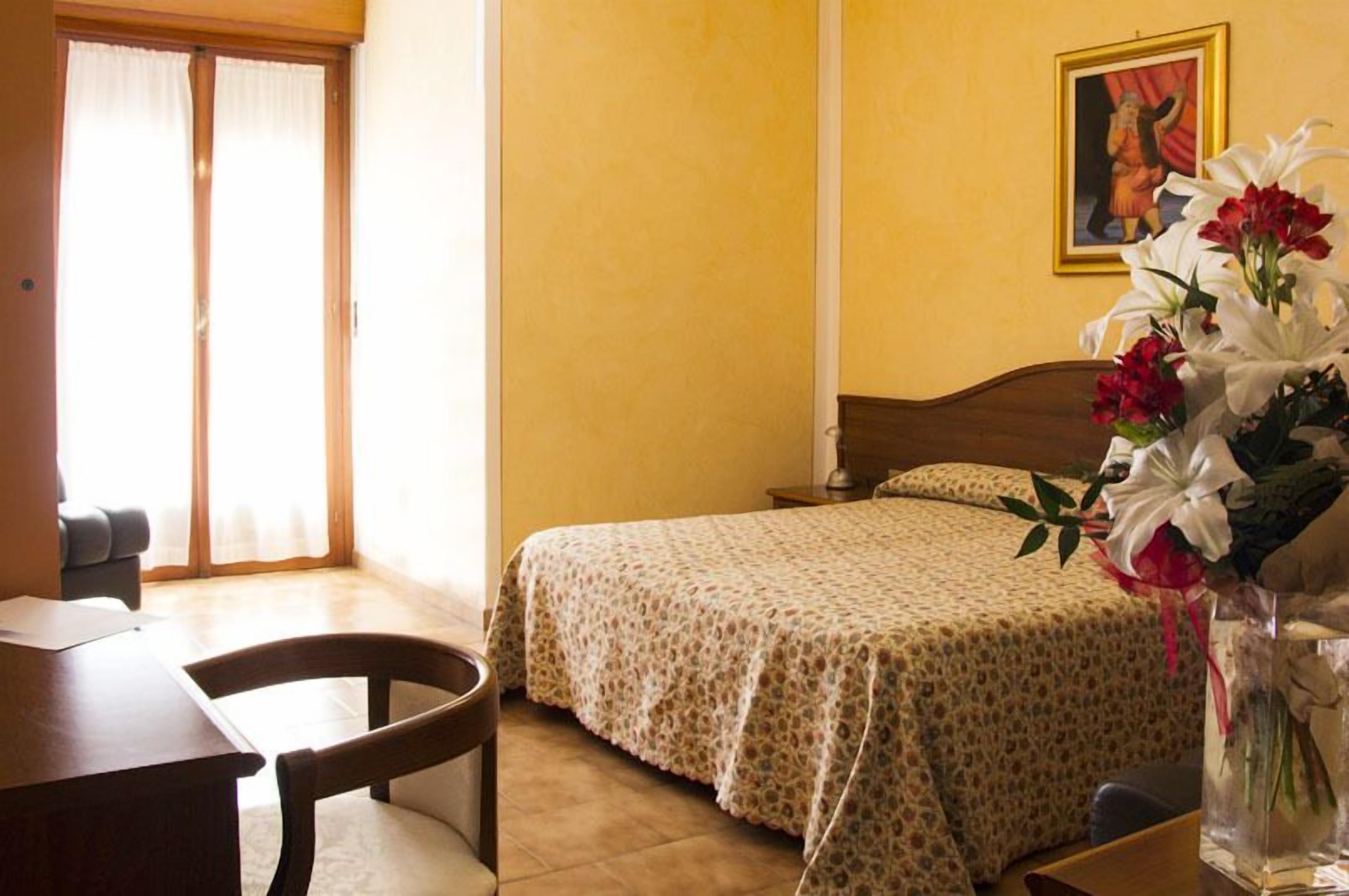 Une chambre d’hôtel avec un lit, un bureau, une chaise, un vase avec des fleurs et un tableau au mur.