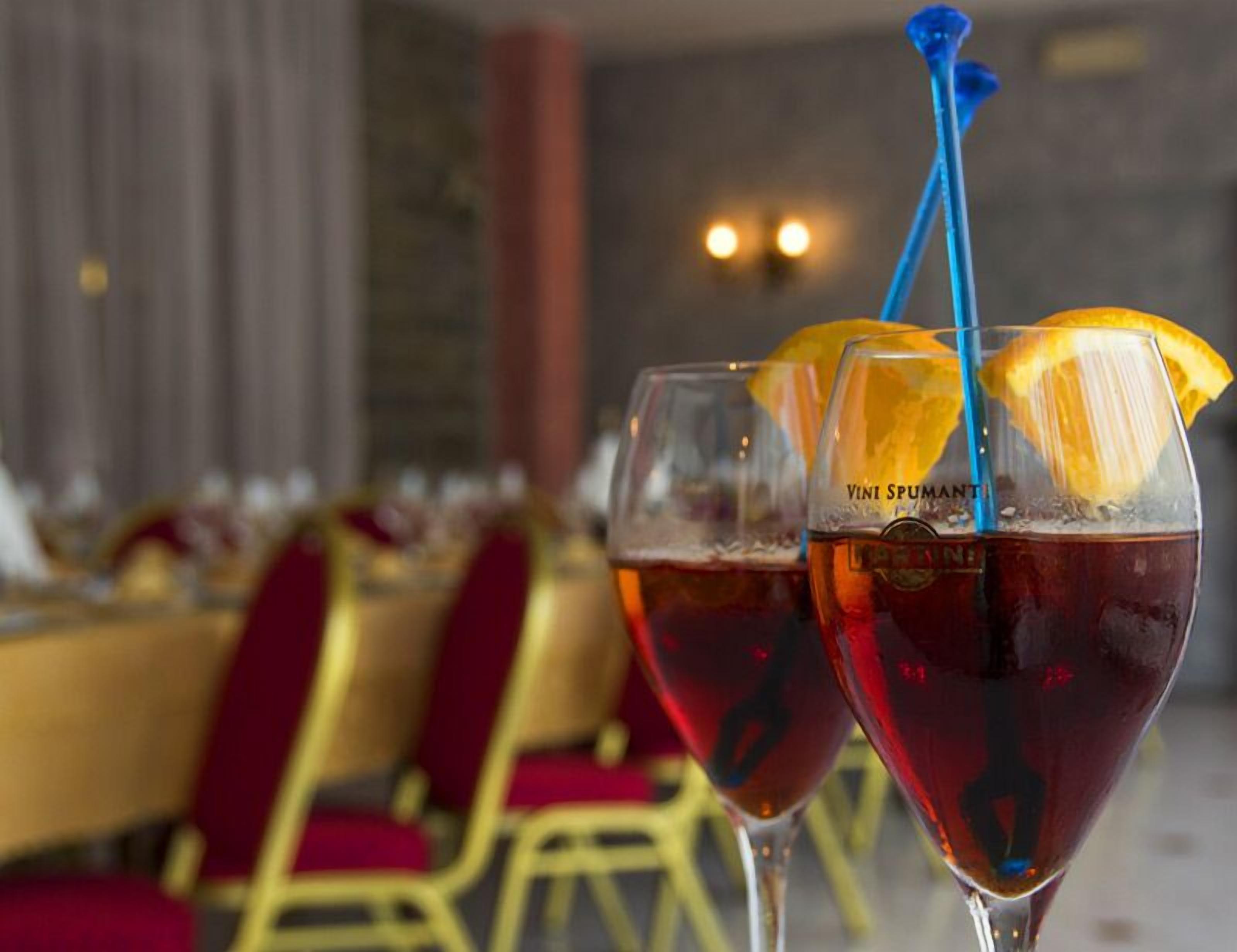 Two glasses of red wine with orange slices and blue straws, placed on a table with a blurred background of a dining area.