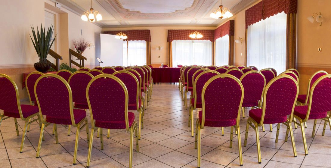 Une salle de conférence avec des rangées de chaises rouges et une longue table, agrémentée d’un lustre et de grandes fenêtres.
