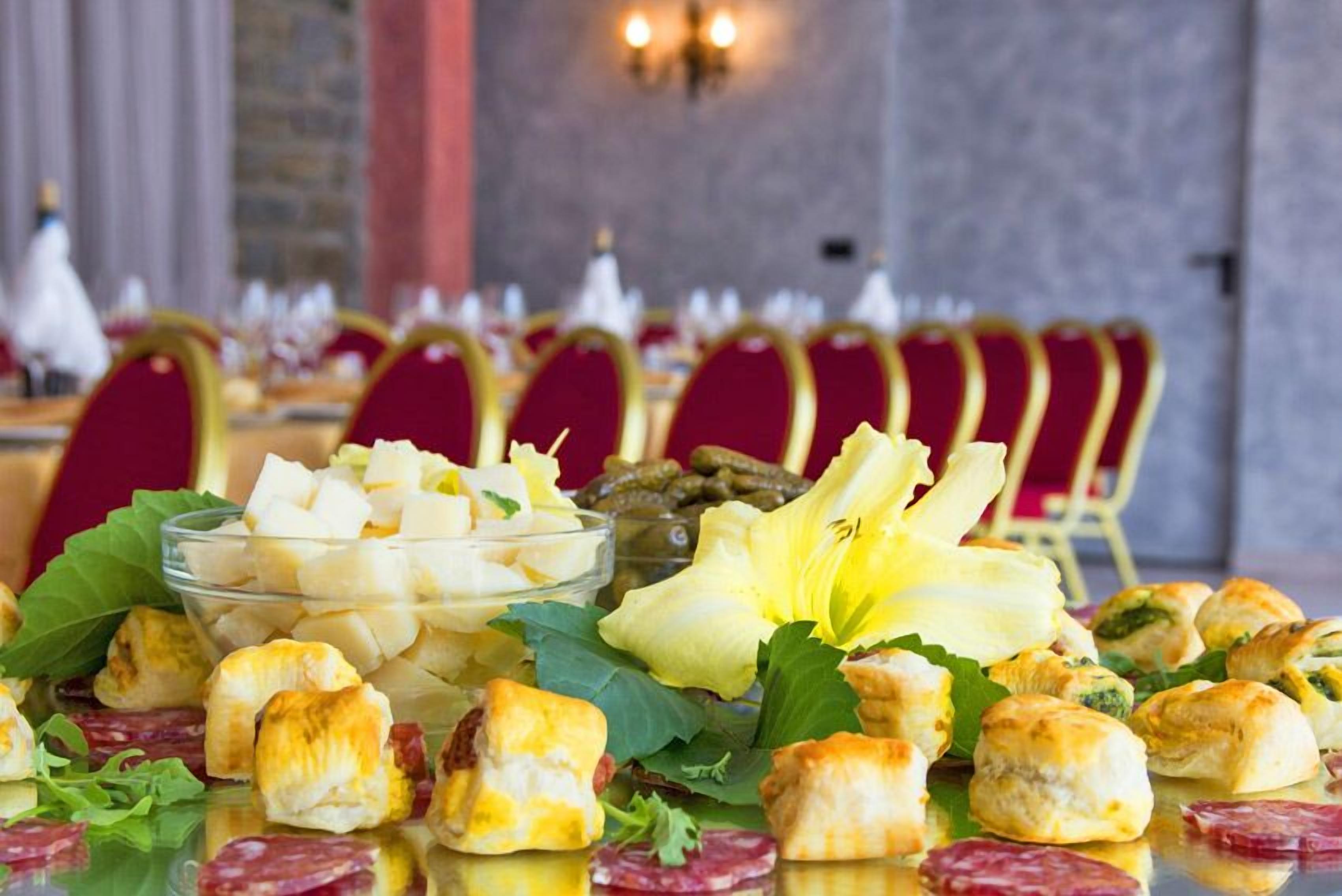 Ein Tisch mit verschiedenen Vorspeisen, darunter gewürfelter Käse, gefüllte Teigtaschen und Blattsalat, vor einer Kulisse aus roten Stühlen und einer Steinsäule.