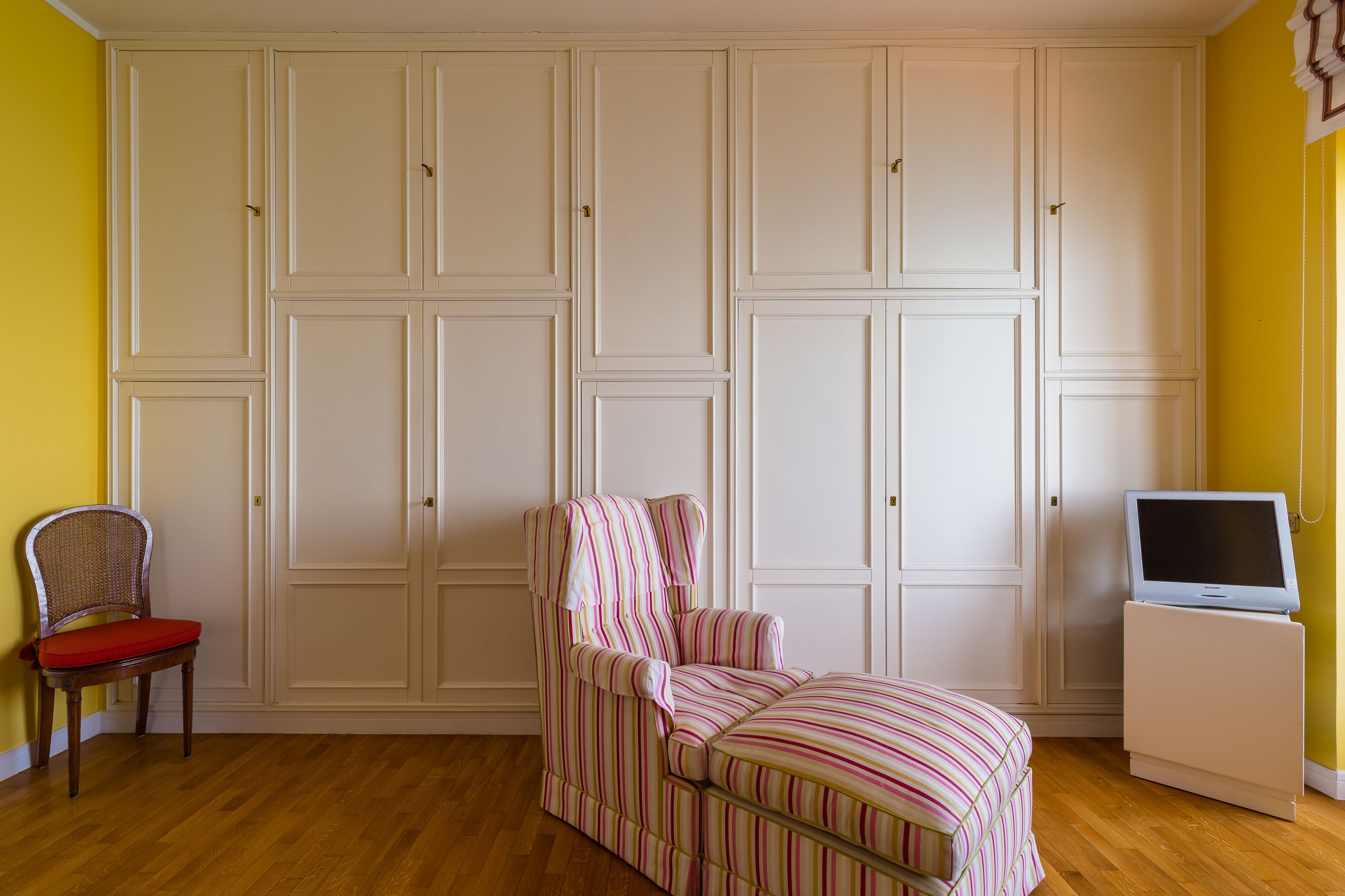A room with a wooden floor, a striped armchair, a red chair, a television on a stand, and a white wardrobe.