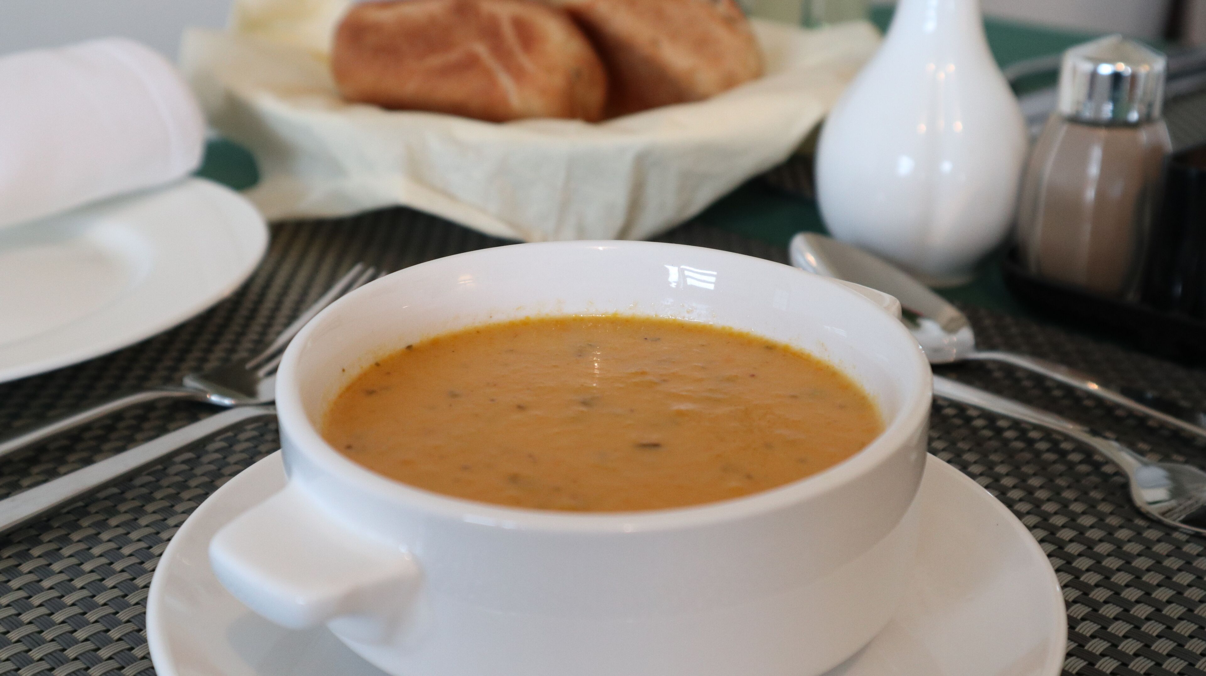 A white bowl of soup on a white saucer, placed on a table with a woven mat.
