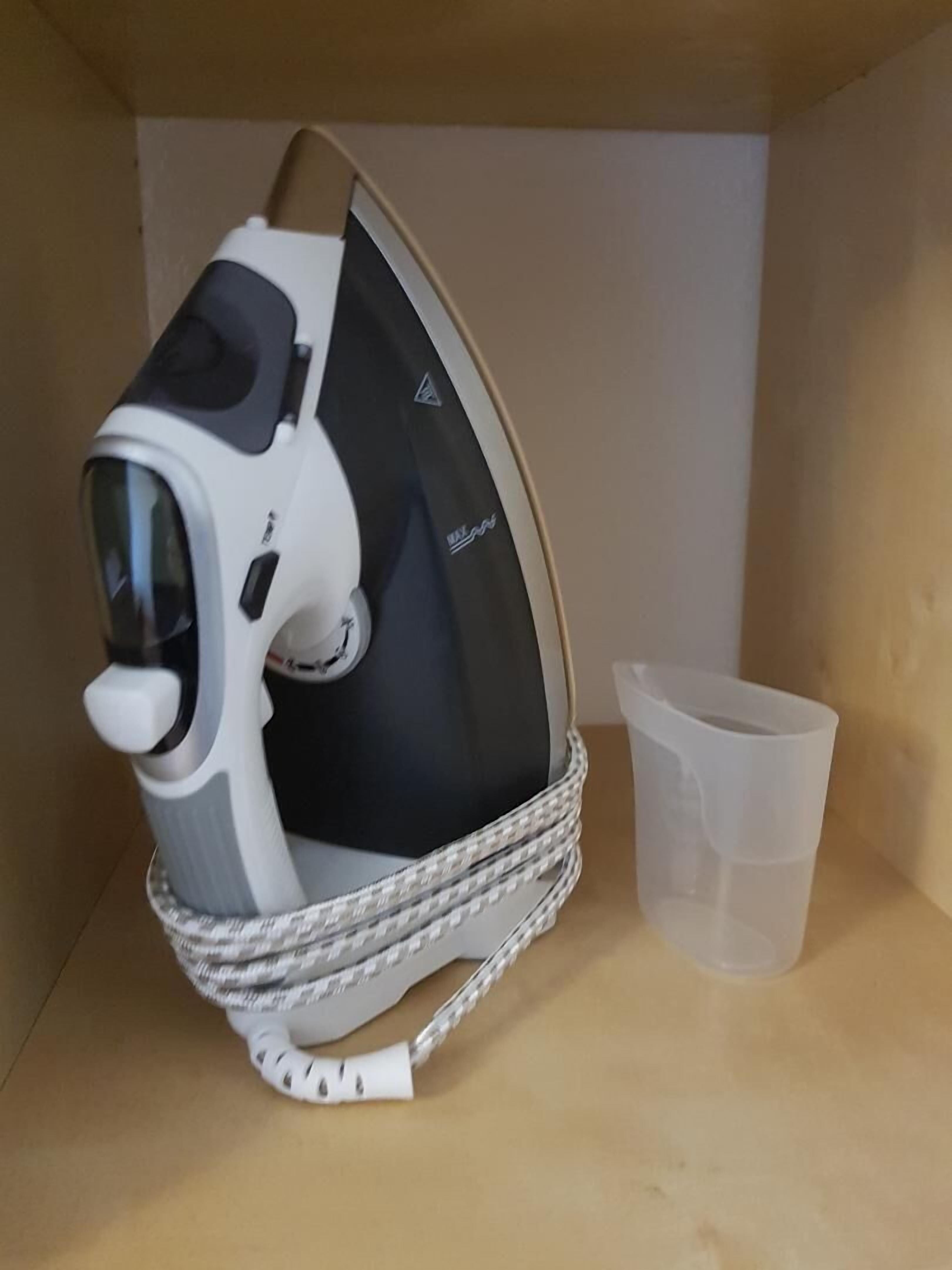 Una plancha compacta y moderna con un panel de control, un cable de alimentación y una taza de plástico sobre una estantería.