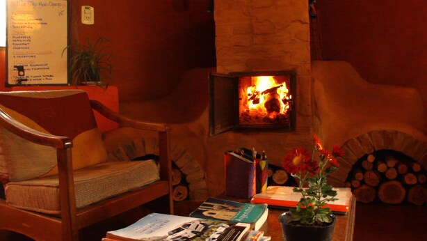 A cozy room with a fireplace, a wooden chair, a table with books and a plant, and a menu board on the wall. - El Pueblito Ecohostel (El Bolson)