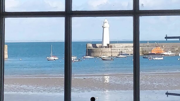 Vista attraverso una finestra su un porto con un faro, barche e una spiaggia sabbiosa.