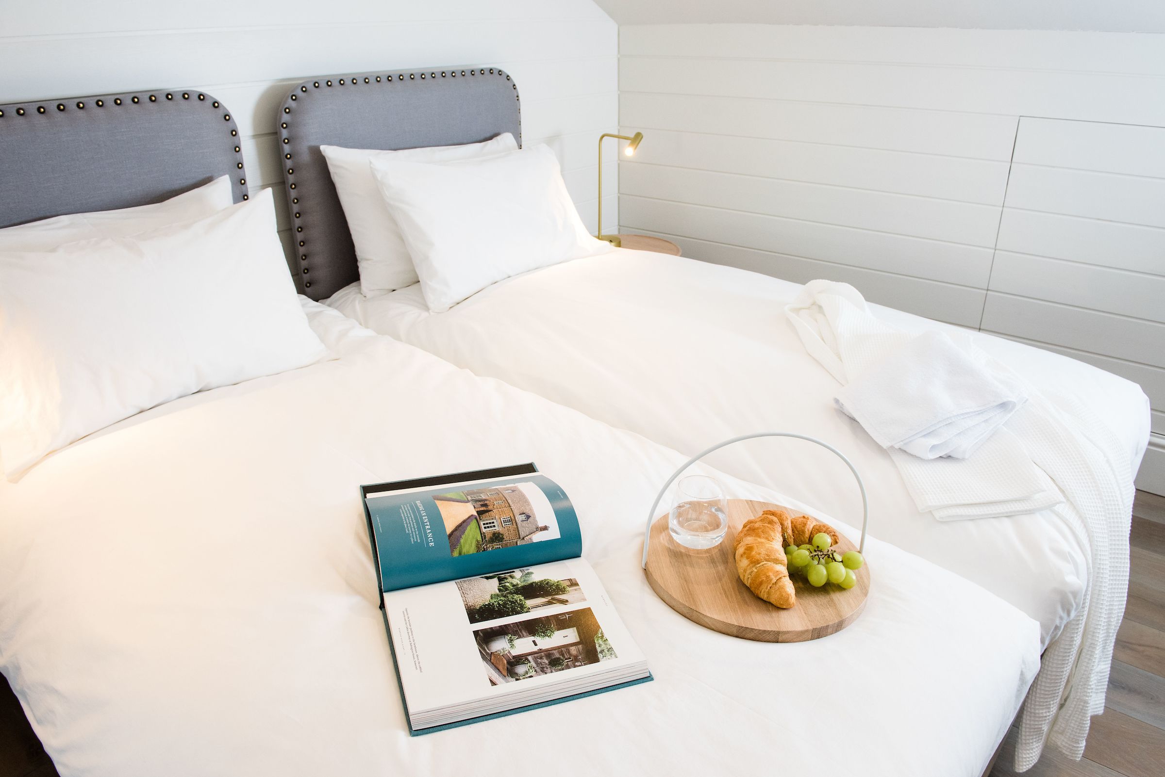 Un lit bien fait, avec du linge de lit blanc, un livre et un plateau en bois contenant des croissants et des raisins.