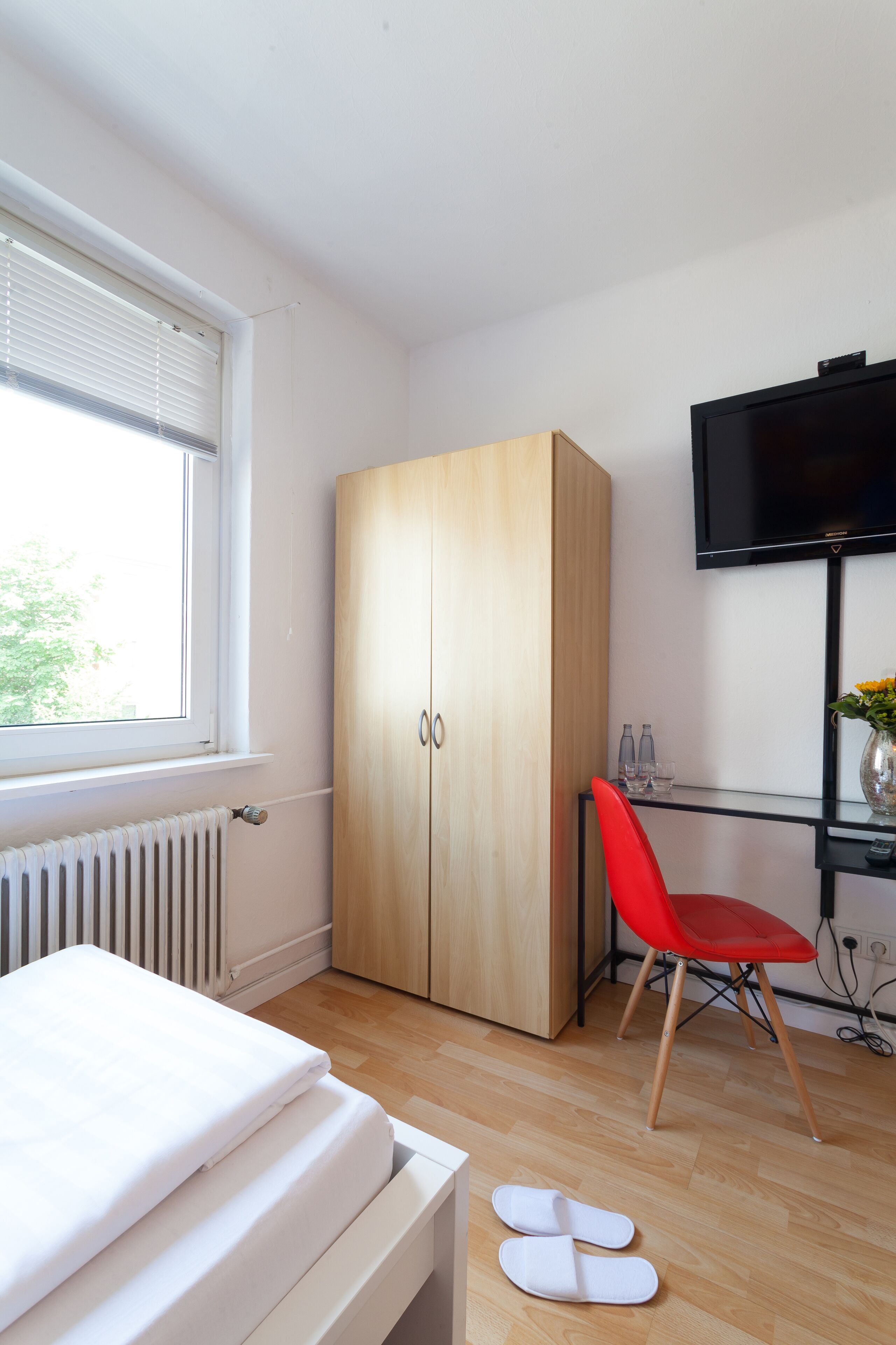 A bedroom with a bed, a wardrobe, a red chair, a TV, and a window with blinds.