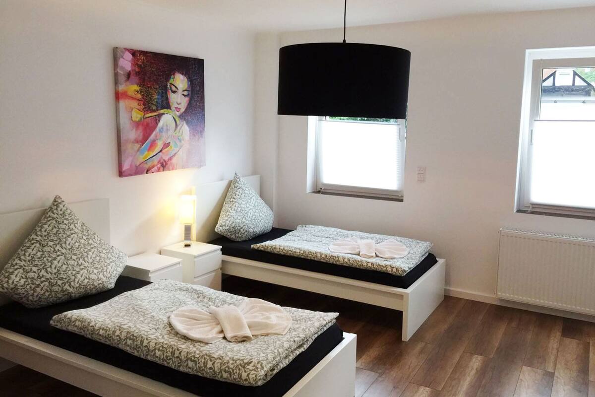 Un dormitorio con dos camas individuales, un cuadro en la pared, una ventana con persianas y una lámpara colgante.