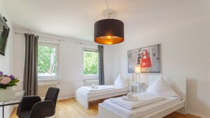 A modern hotel room with two single beds, a desk with a chair, a large window with curtains, a radiator, and a painting on the wall. - City Apartment Hannover (Hannover)
