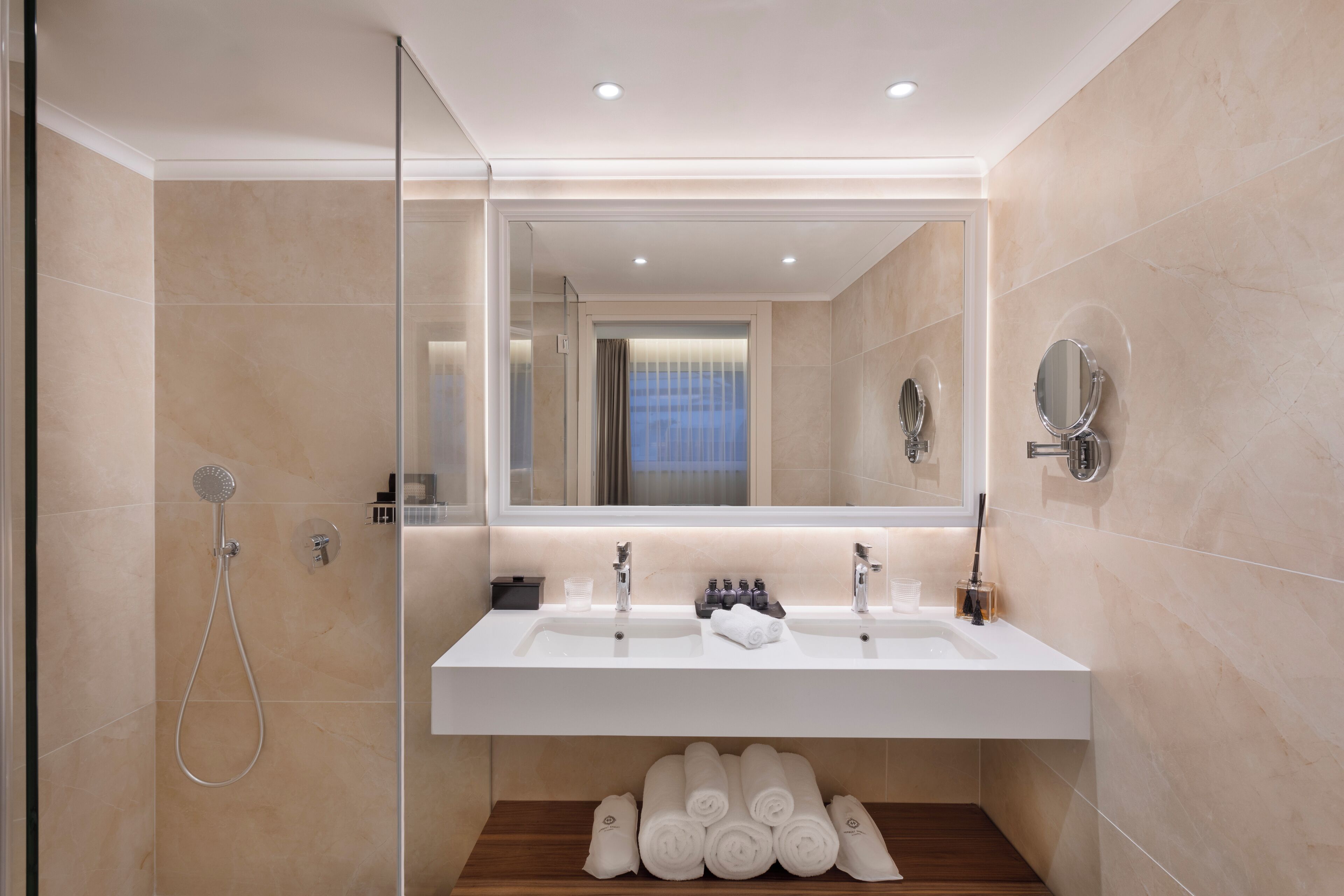 Un baño moderno con un espejo grande, dos lavamanos, una ducha y toallas ordenadas en un estante.