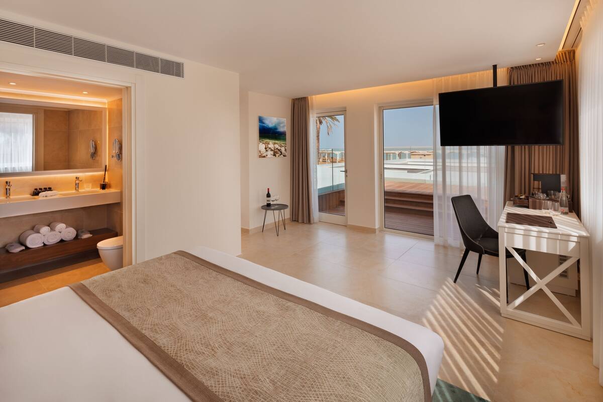 A modern hotel room with a large bed, a desk with a chair, a flat-screen TV, and a balcony with a view of the beach.