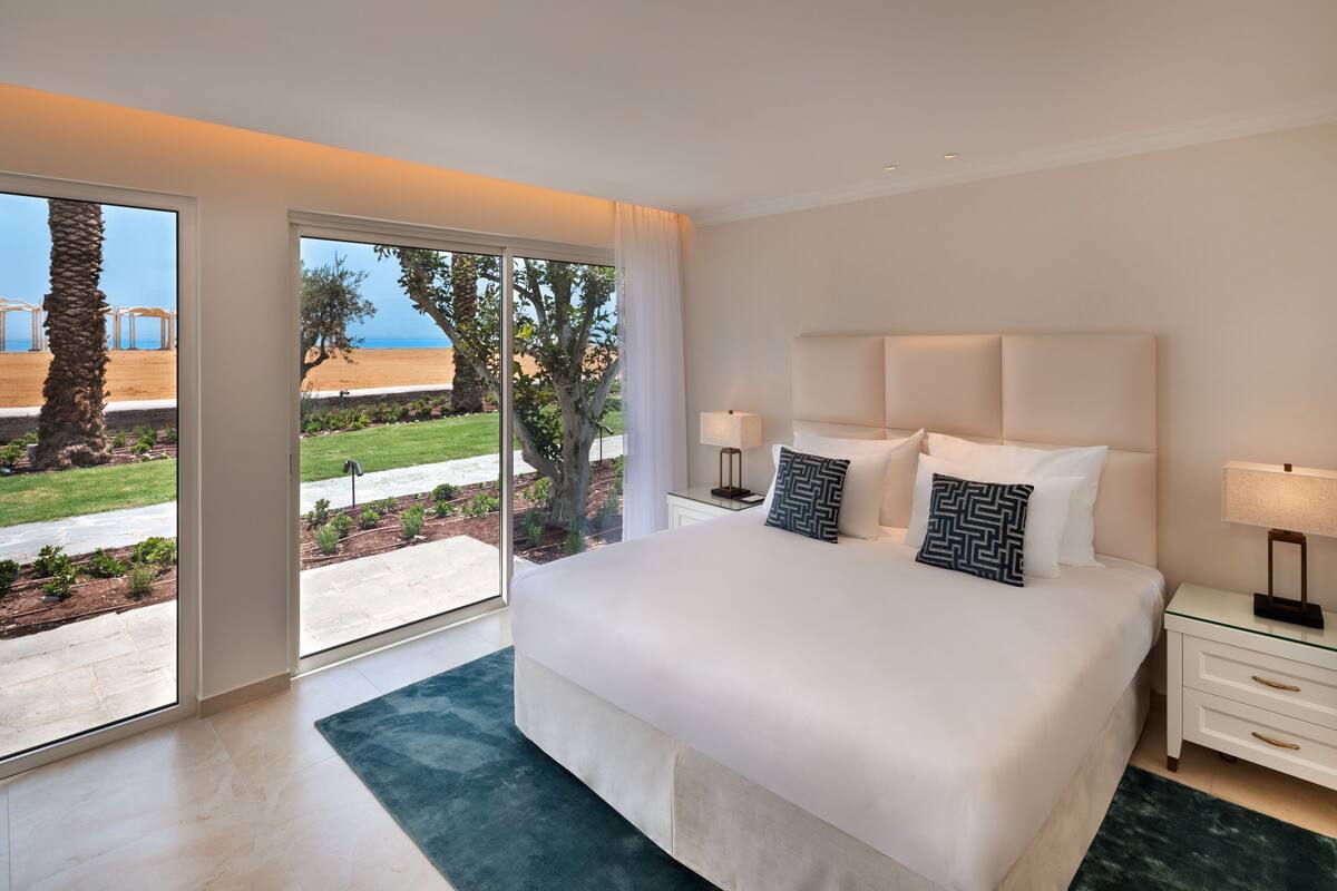 A modern hotel room with a large bed, two bedside tables, and a view of a beach and palm trees.