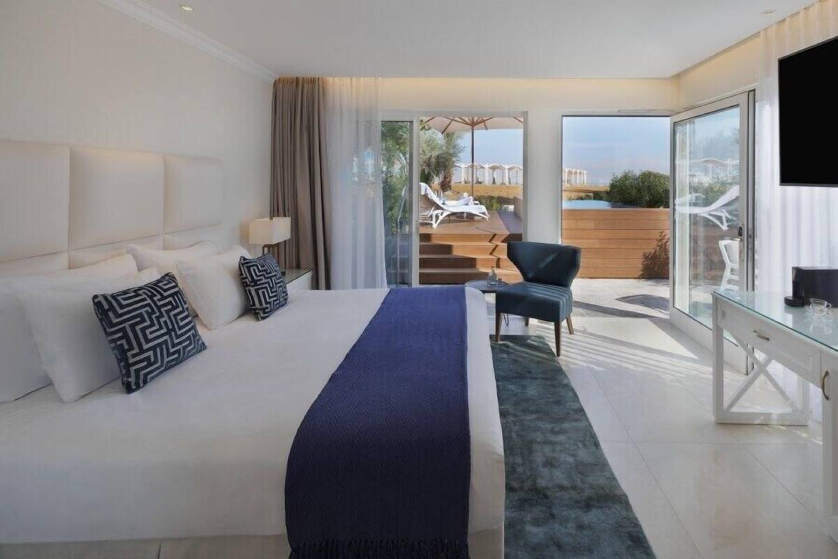 A modern hotel room with a large bed, a glass door leading to a balcony with outdoor seating, and a TV on the right side.