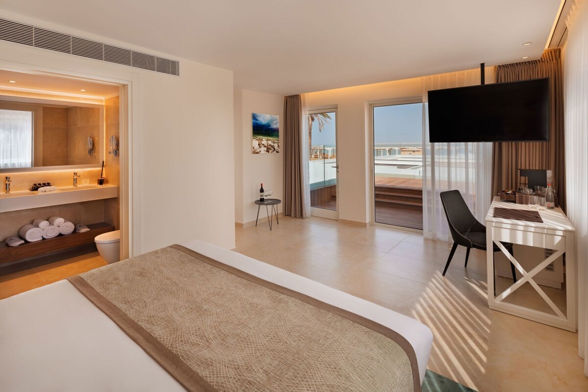 A hotel room with a bed, a desk with a TV, a bathroom with a mirror, and a view of the sea.