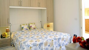 Un dormitorio con una cama, dos mesitas de noche, una mesita auxiliar, una ventana y una puerta.
