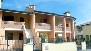 Un edificio de dos pisos con un balcón en el nivel superior, una escalera que conduce a la entrada y un camino de entrada con portón.