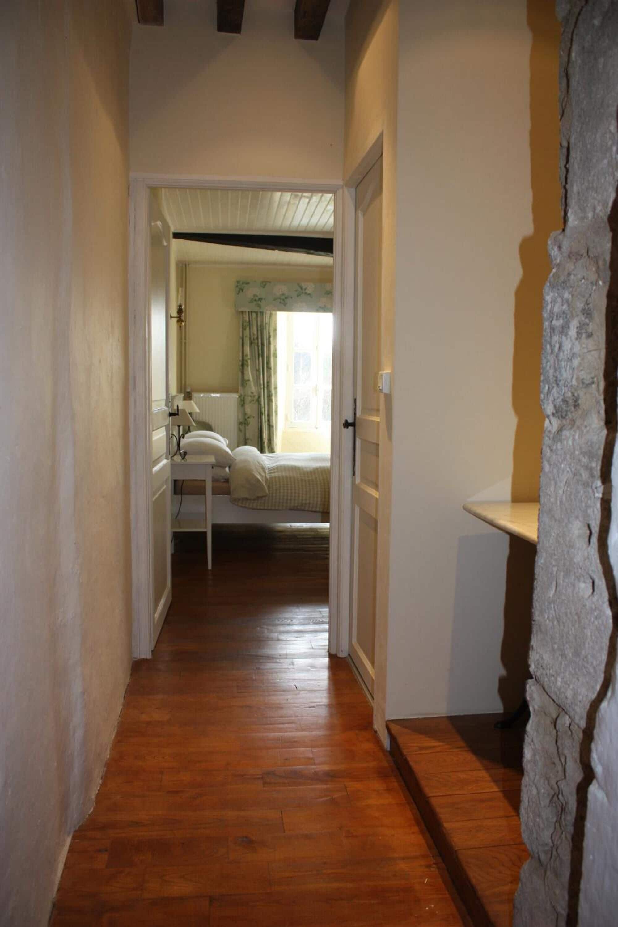 A narrow hallway with wooden flooring leading to a bedroom with a bed, a bedside table, and a window with curtains.