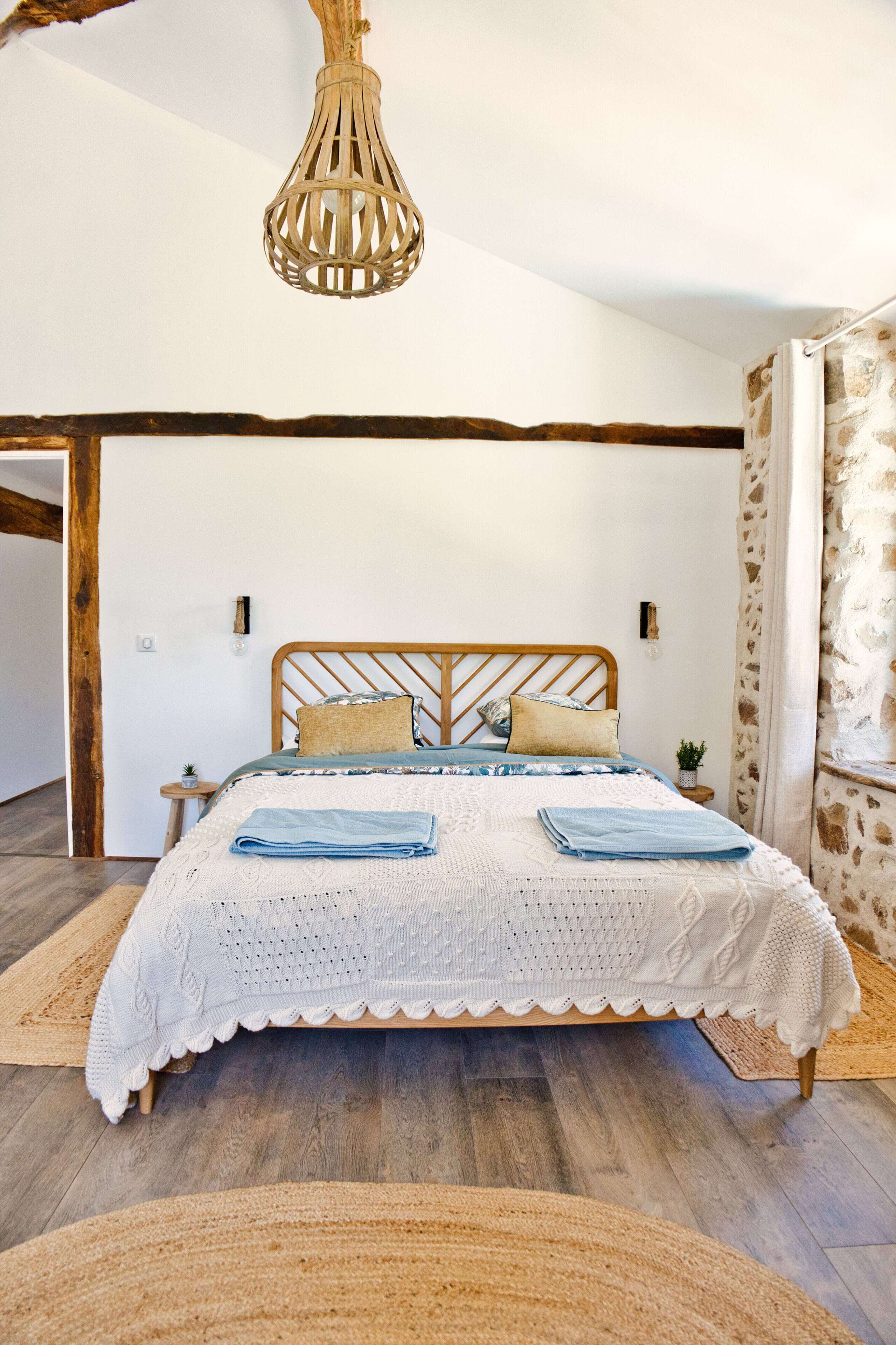 A bedroom with a wooden bed, white bedding, a hanging light fixture, and a stone wall.