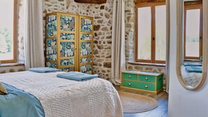A bedroom with a stone wall, wooden beams, a bed with a white and blue bedspread, a green chest of drawers, and a large mirror. - Les Picardies (Lesterps)