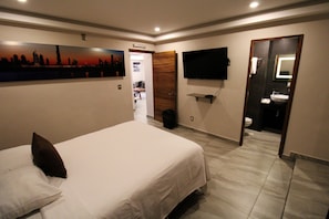 A hotel room with a bed, a TV mounted on the wall, a bathroom with a sink and mirror, and a cityscape painting on the wall. - Oslo 10 (Mexico City)
