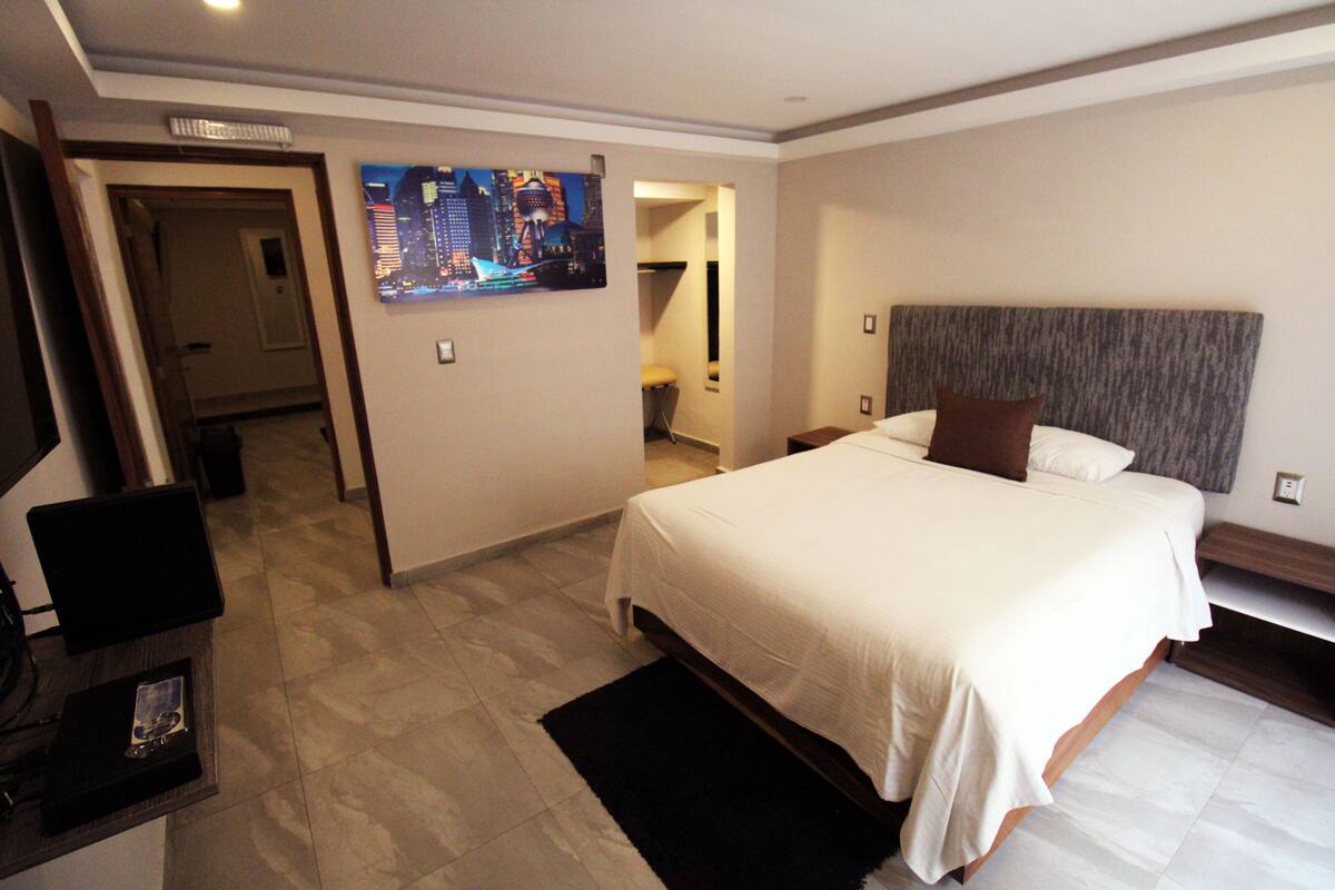 Une chambre d’hôtel dotée d’un grand lit, d’un téléviseur à écran plat, d’un bureau avec un ordinateur et d’un tableau représentant un paysage urbain accroché au mur.