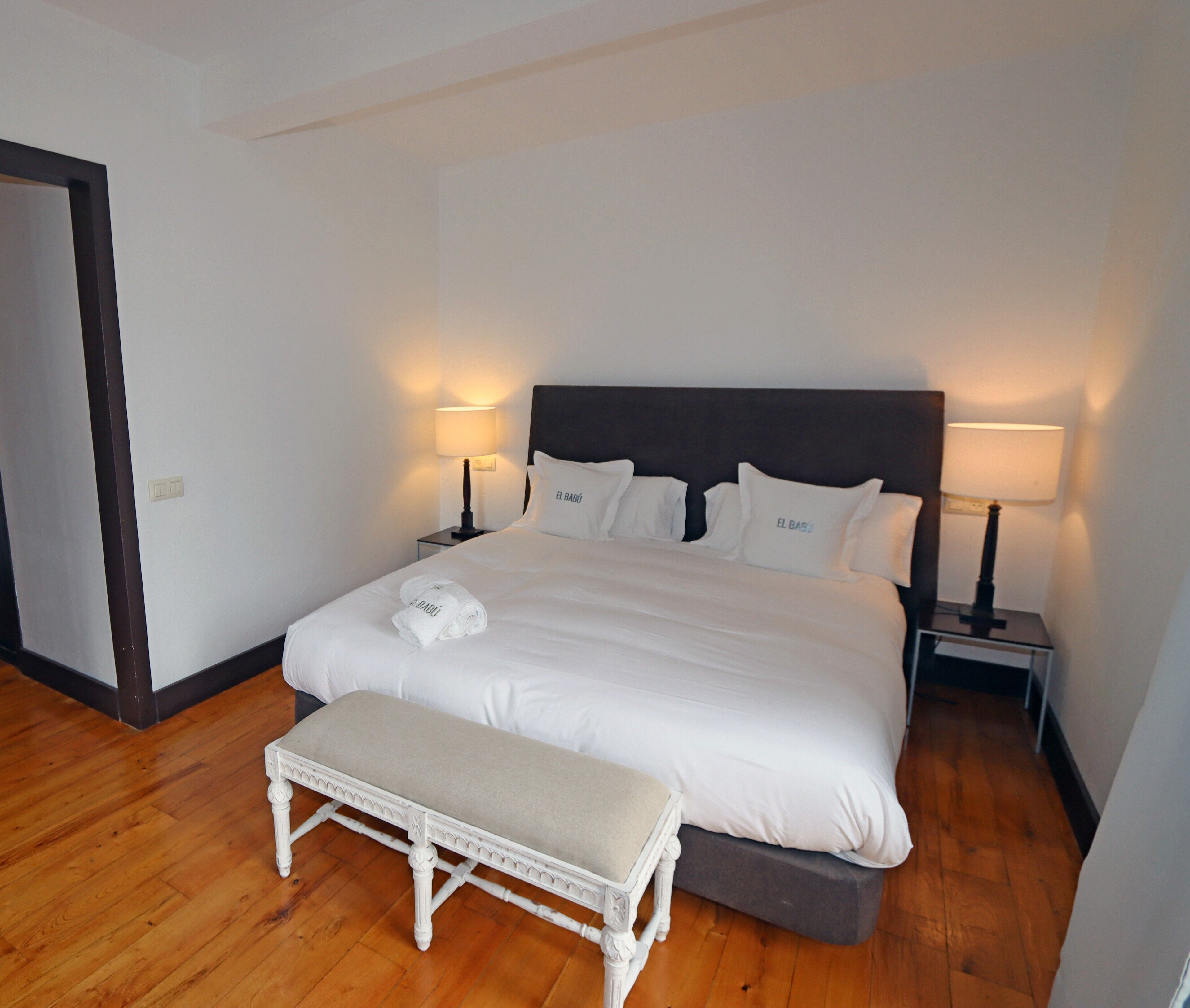 A neatly made bed with white linens and pillows, two bedside lamps, a wooden bench, and a door to the left.