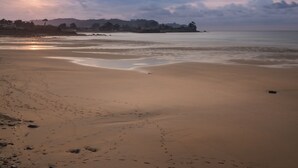 Una playa con huellas que conducen al agua, un mar tranquilo y una puesta de sol al fondo.