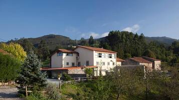 A two-story building with a tiled roof, surrounded by greenery and hills in the background.