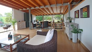 Un salón amplio con techo de madera, un sofá de mimbre, una mesa de centro de vidrio y un comedor con sillas y una mesa.