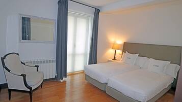 A hotel room with a double bed, a chair, a lamp, a window with curtains, and a radiator.