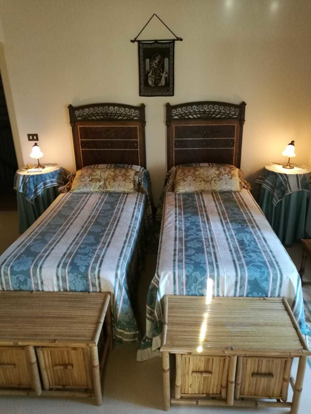 A room with two wooden beds, each with a headboard and a bedside table with a lamp. There is a framed picture on the wall above the beds.