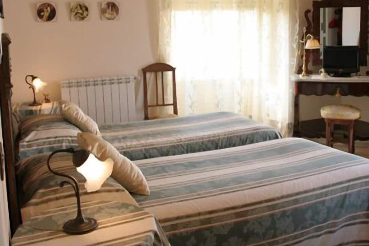 A bedroom with two beds, a wooden ceiling, a desk with a chair, and a window with a curtain.