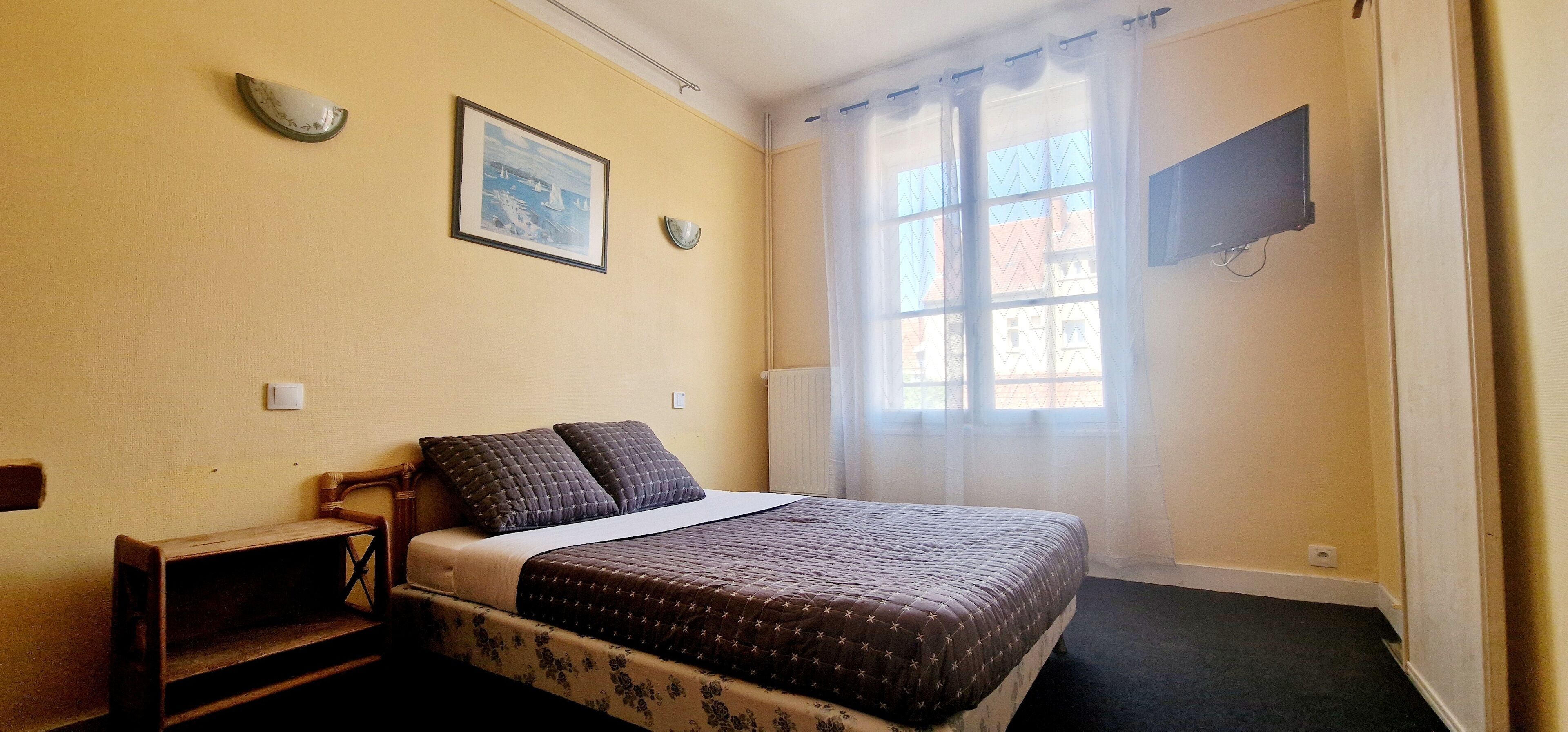 A bedroom with a bed, a bench, a wall-mounted TV, and a window with curtains.
