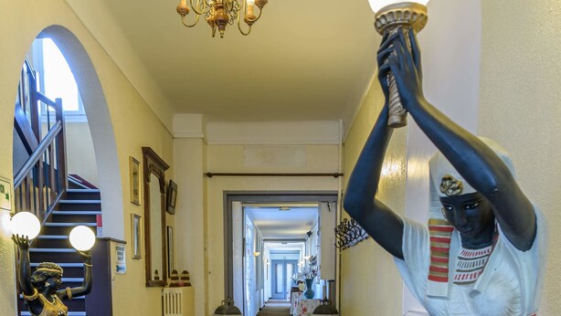 Un pasillo con una estatua de una figura sosteniendo una antorcha, una escalera y un candelabro.