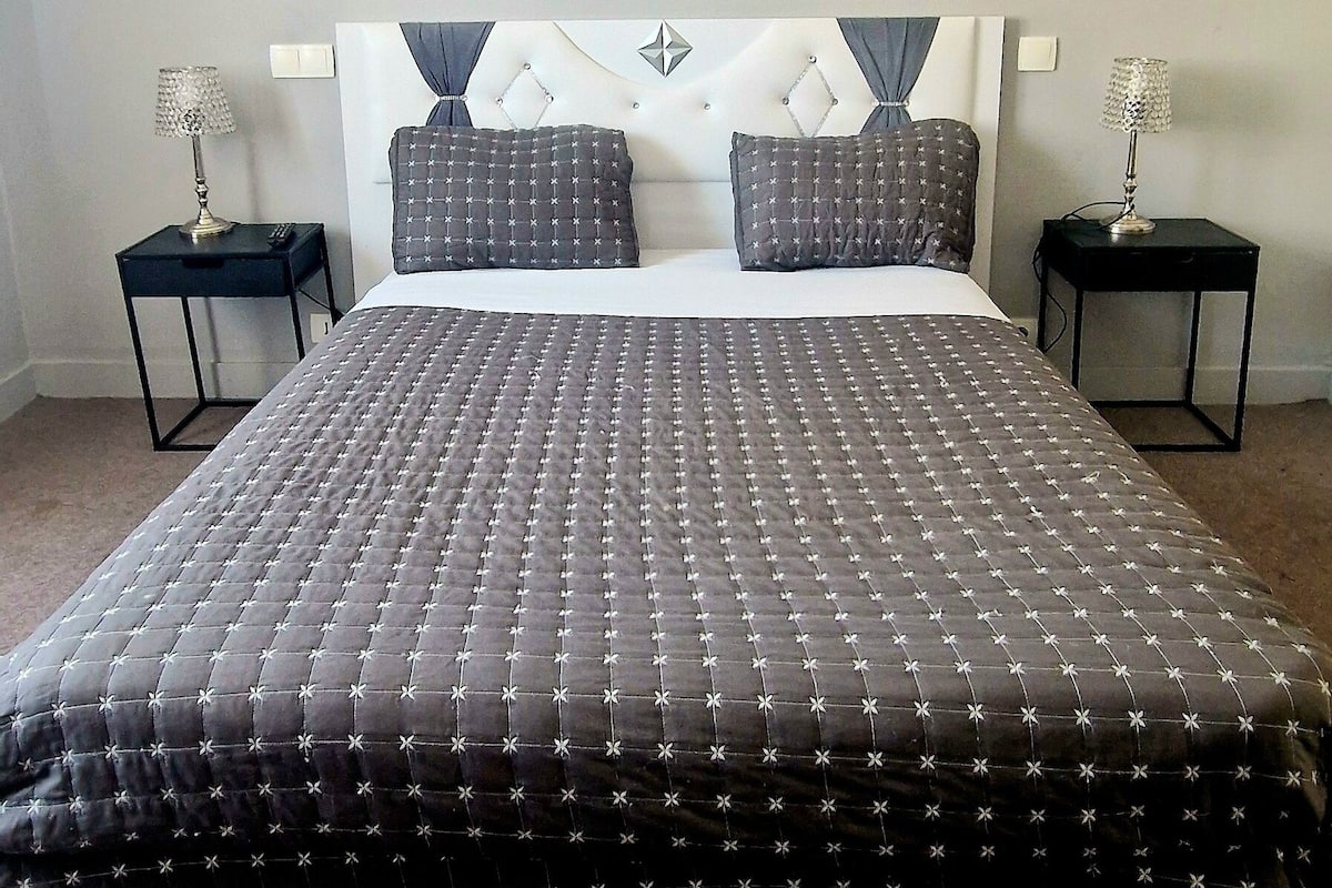 Une chambre d’hôtel avec un grand lit, deux tables de chevet, une tête de lit ornée d’un motif décoratif et un tableau abstrait accroché au mur.