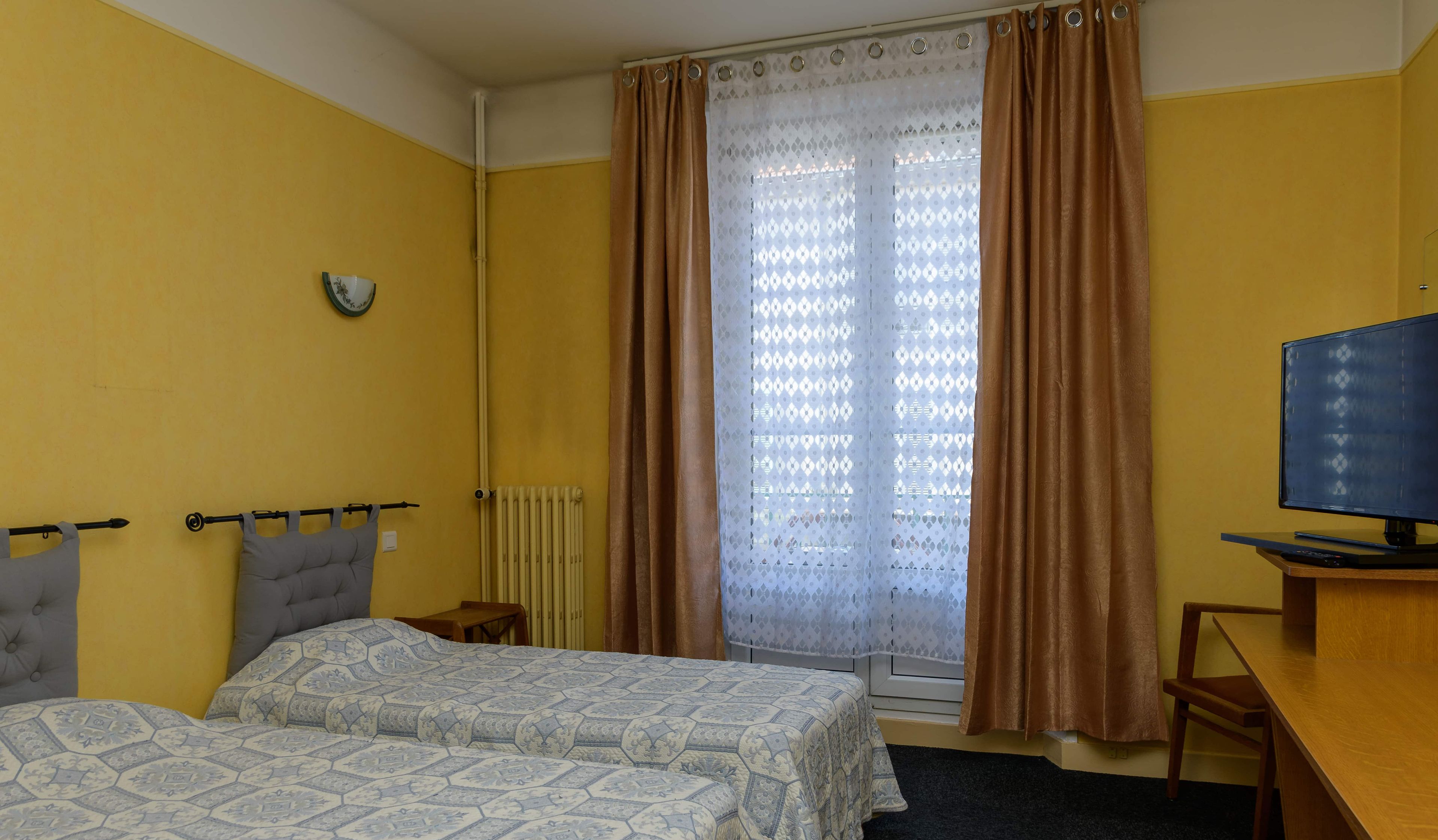 Una habitación de hotel con dos camas, un escritorio con televisor, una ventana con cortinas marrones y un radiador.