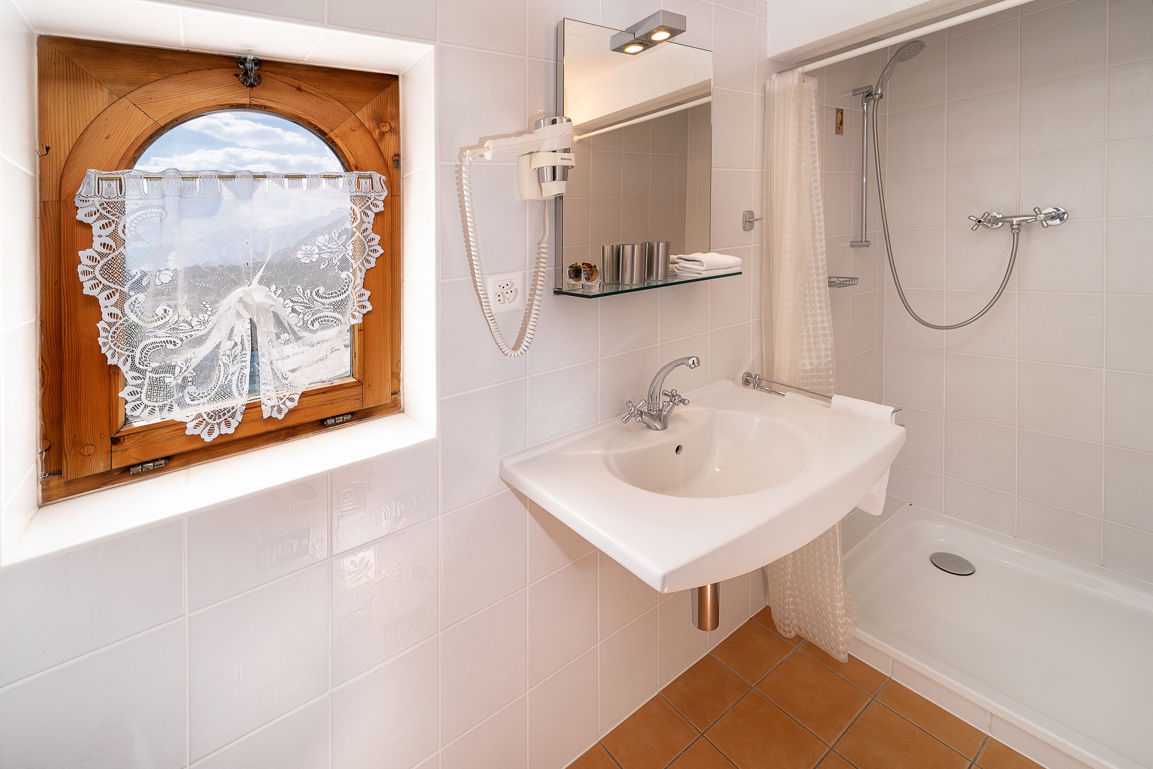A bathroom with a white sink, a mirror, a shower, and a window with a wooden frame and lace curtain.
