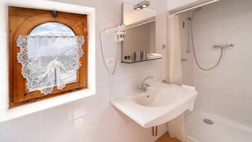 A bathroom with a white sink, a mirror, a shower, and a window with a wooden frame and lace curtain.