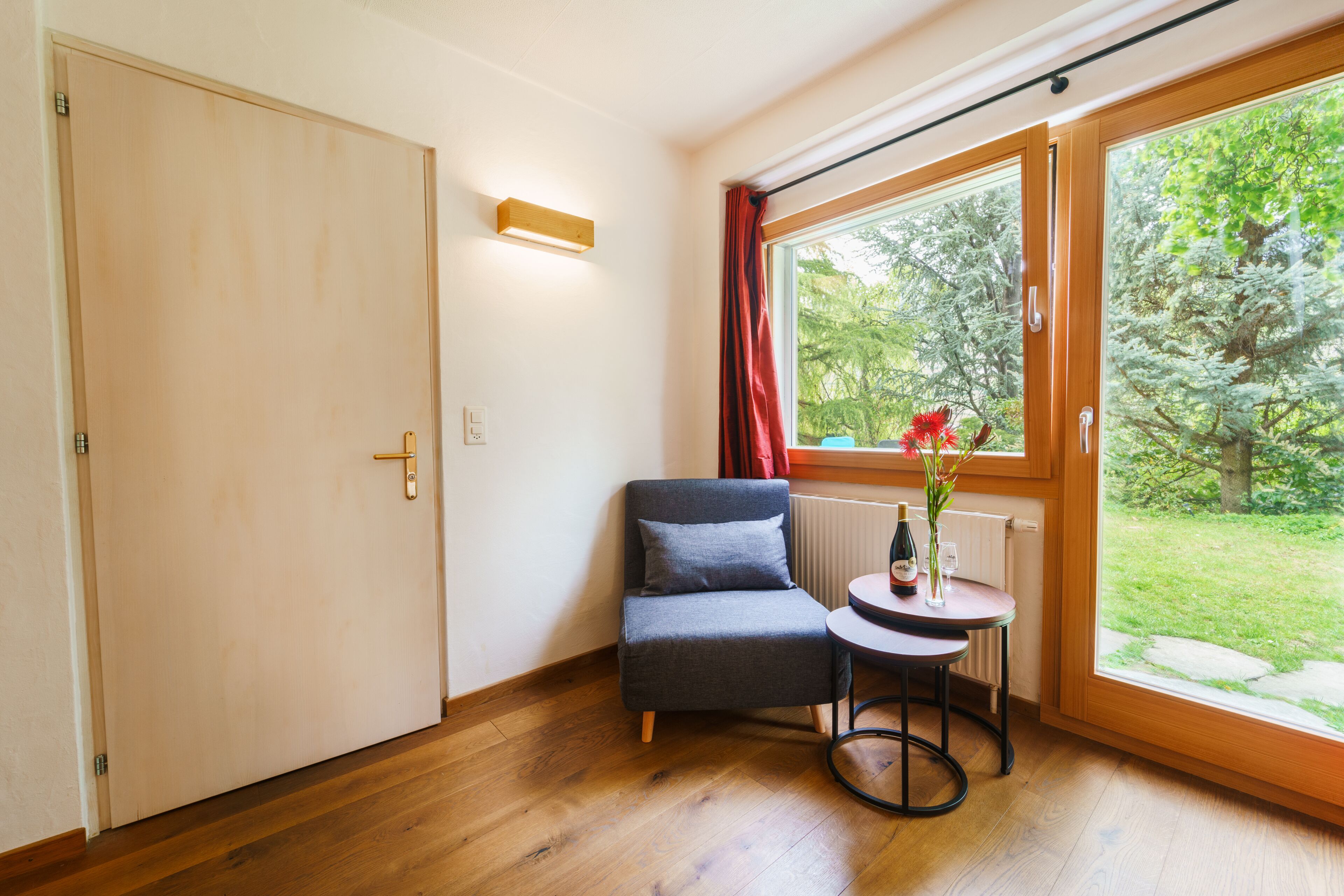 A room with a wooden floor, a grey chair, a small round table with a bottle and flowers, and a window with a view of greenery.