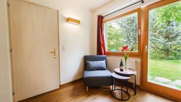 A room with a wooden floor, a grey chair, a small round table with a bottle and flowers, and a window with a view of greenery.