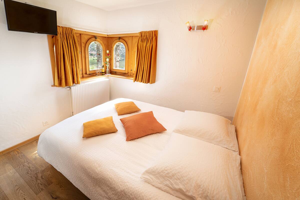 A bedroom with a bed, two windows with curtains, a radiator, and a wall-mounted TV.