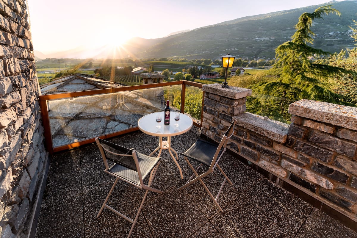 A glass-enclosed patio with a table and chairs, overlooking a scenic valley with a sunset.