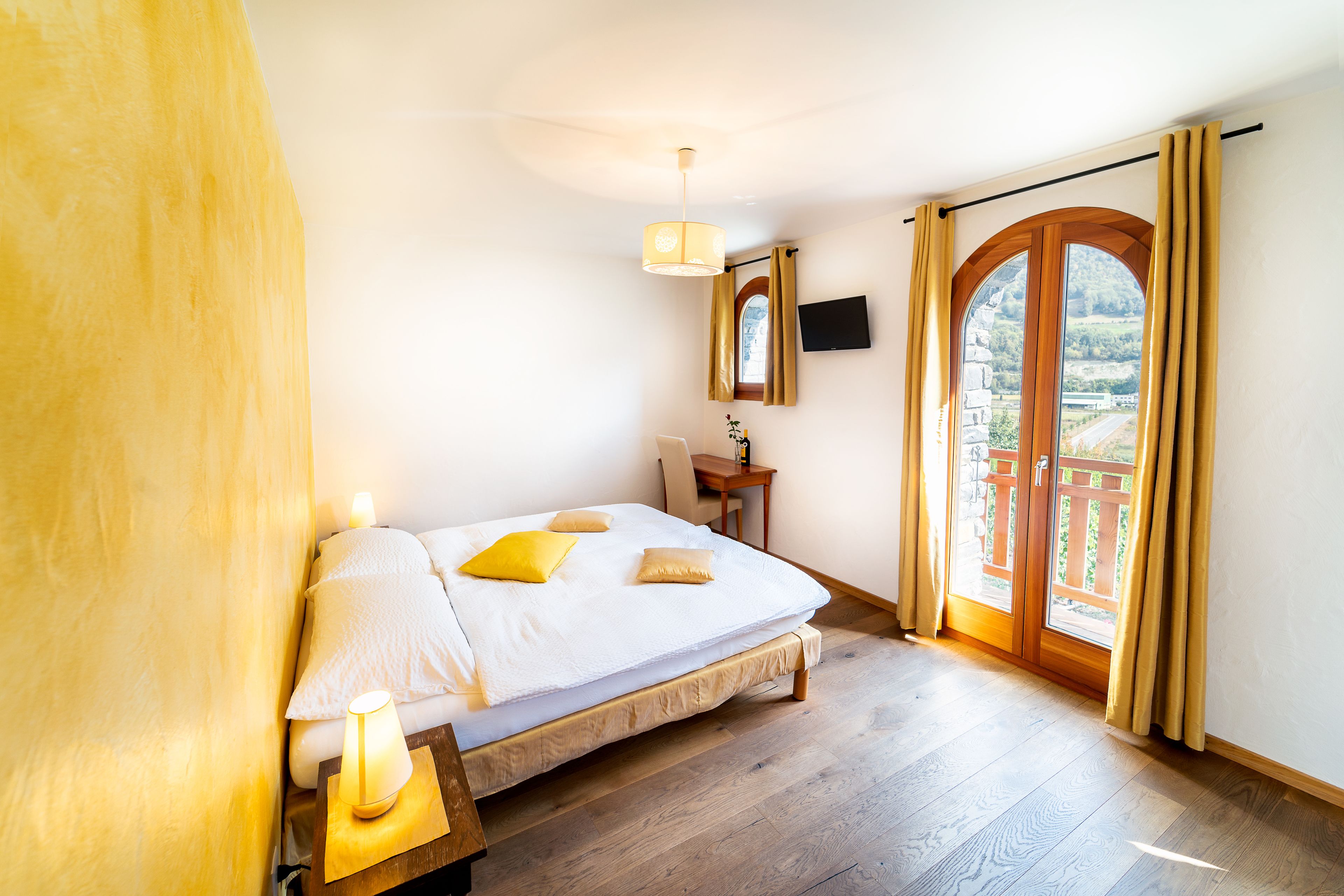 Quarto com cama, luminária de cabeceira, piso de madeira e vista para uma varanda com guarda-corpo de madeira.